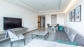 A modern living room with a flat-screen TV, a grey sofa, a white armchair, a coffee table, and a dining area with a table and chairs.