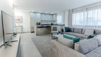 Un salon moderne avec un canapé gris, un téléviseur à écran plat et un coin cuisine doté de meubles de couleur claire et d’un tabouret de bar.