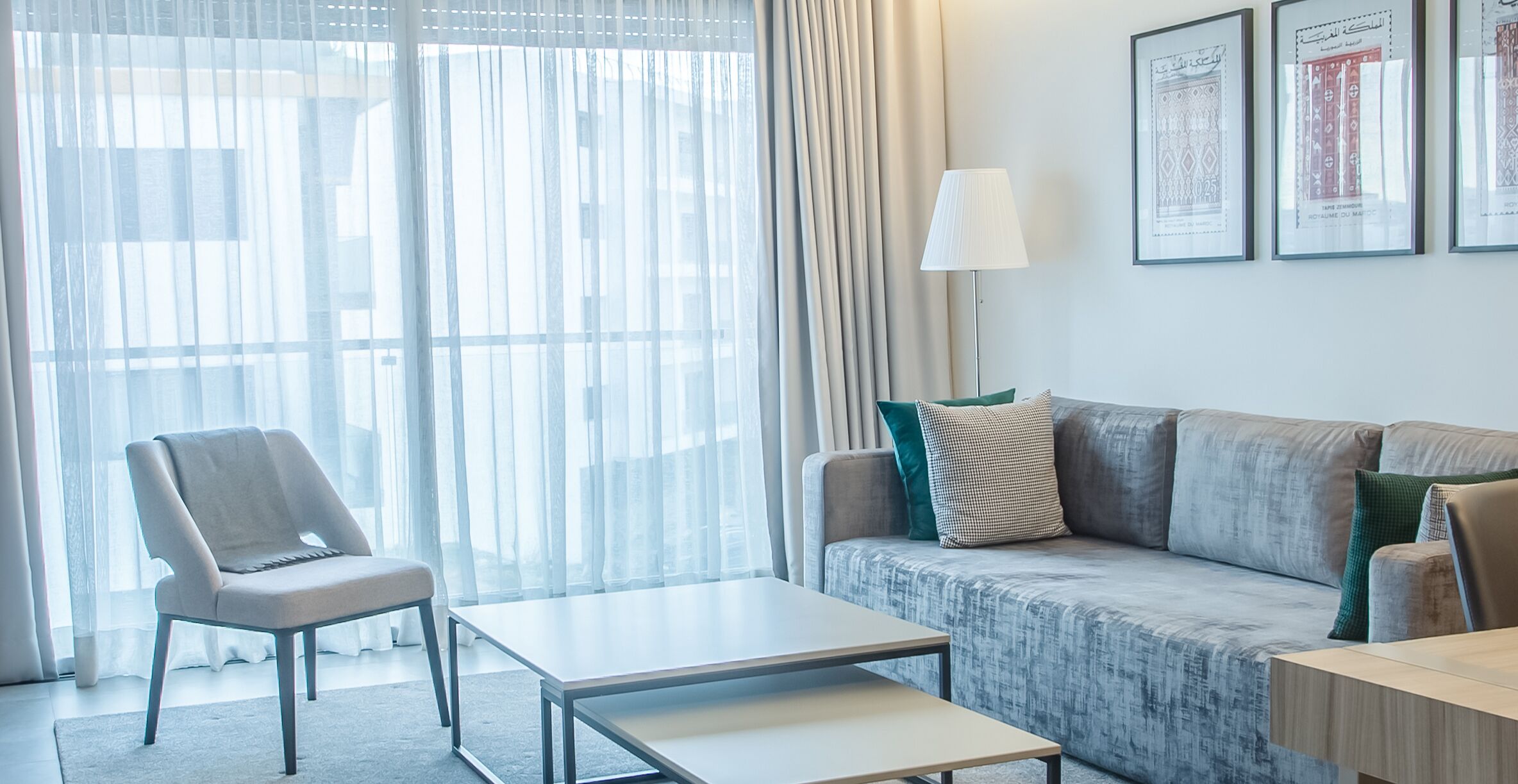 Een moderne woonkamer met een grijze bank, een witte tafel en een grijze fauteuil. Er hangen afbeeldingen in lijsten aan de muur en op een bijzettafeltje staat een lamp.