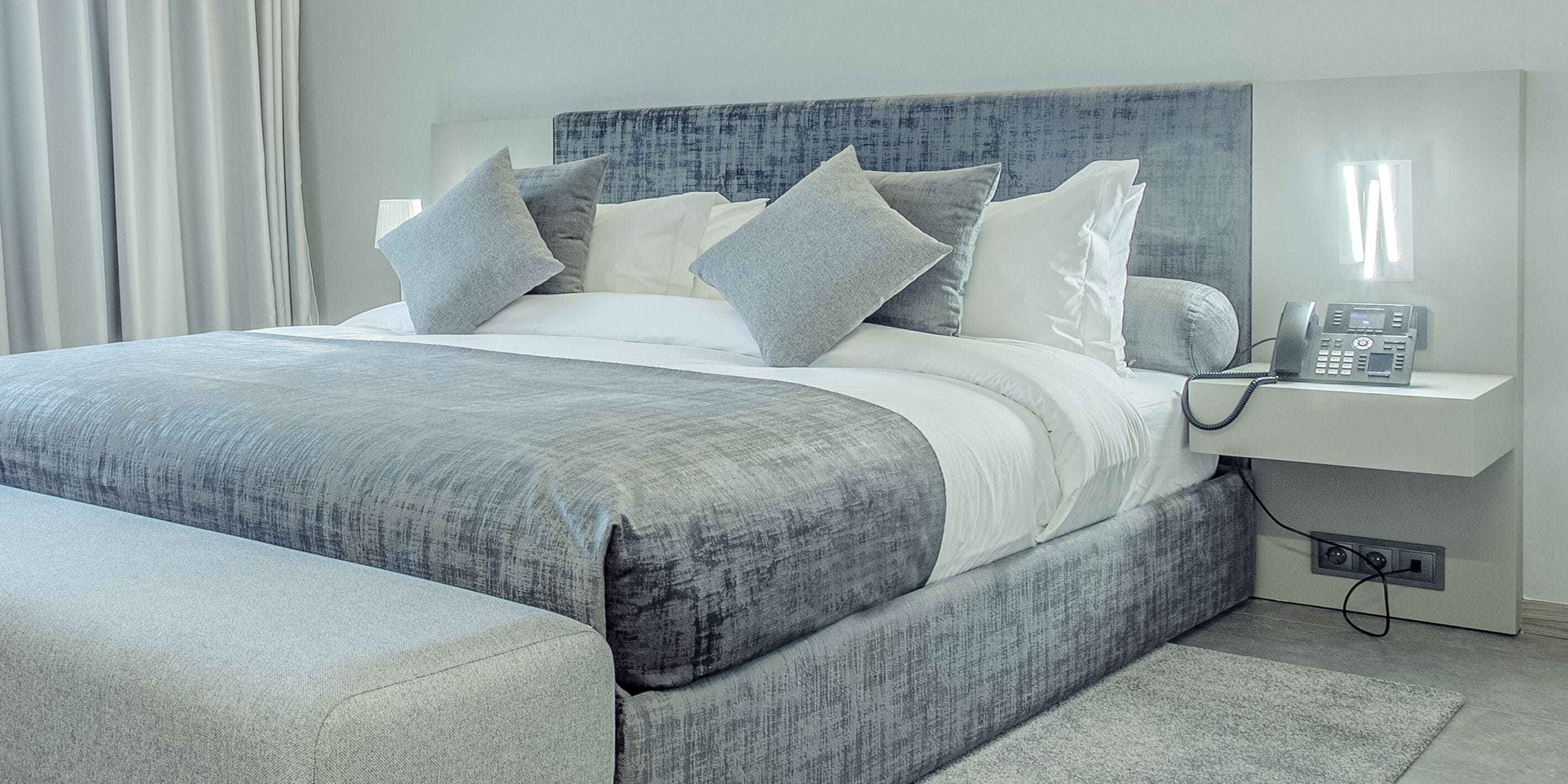 A modern bedroom with a large bed, grey bedding, and a bedside phone.