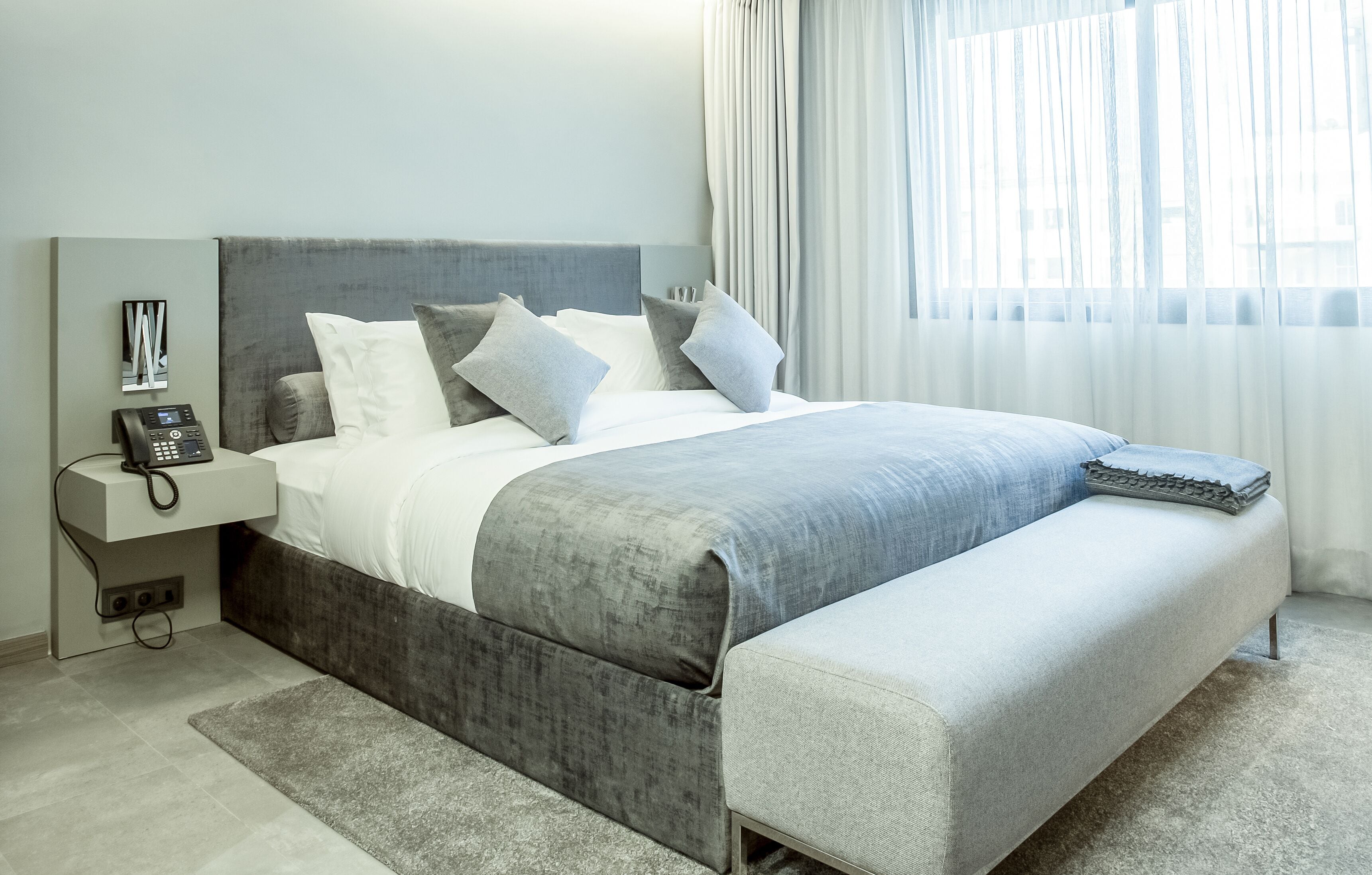 A modern bedroom with a grey upholstered bed, a bedside phone, and a large window with sheer curtains.
