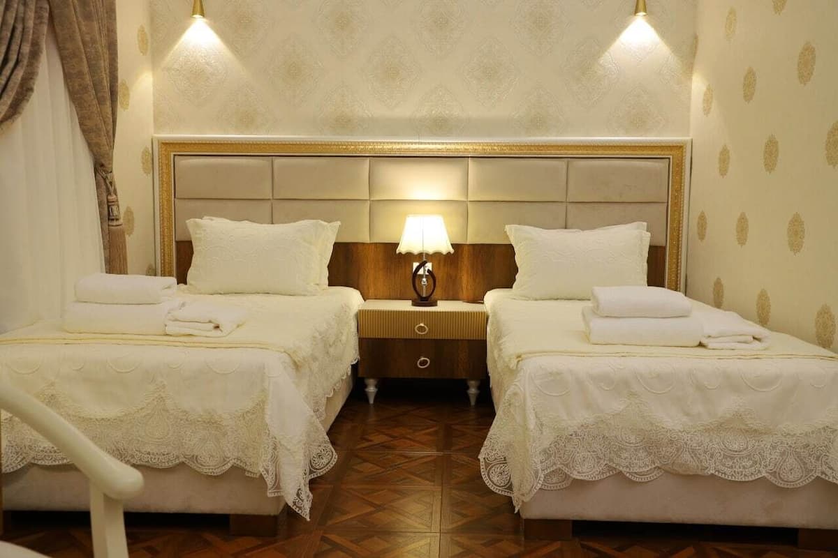 A hotel room with two beds, each with a white bedspread and matching pillows, a wooden nightstand with a lamp, and a patterned wallpaper.