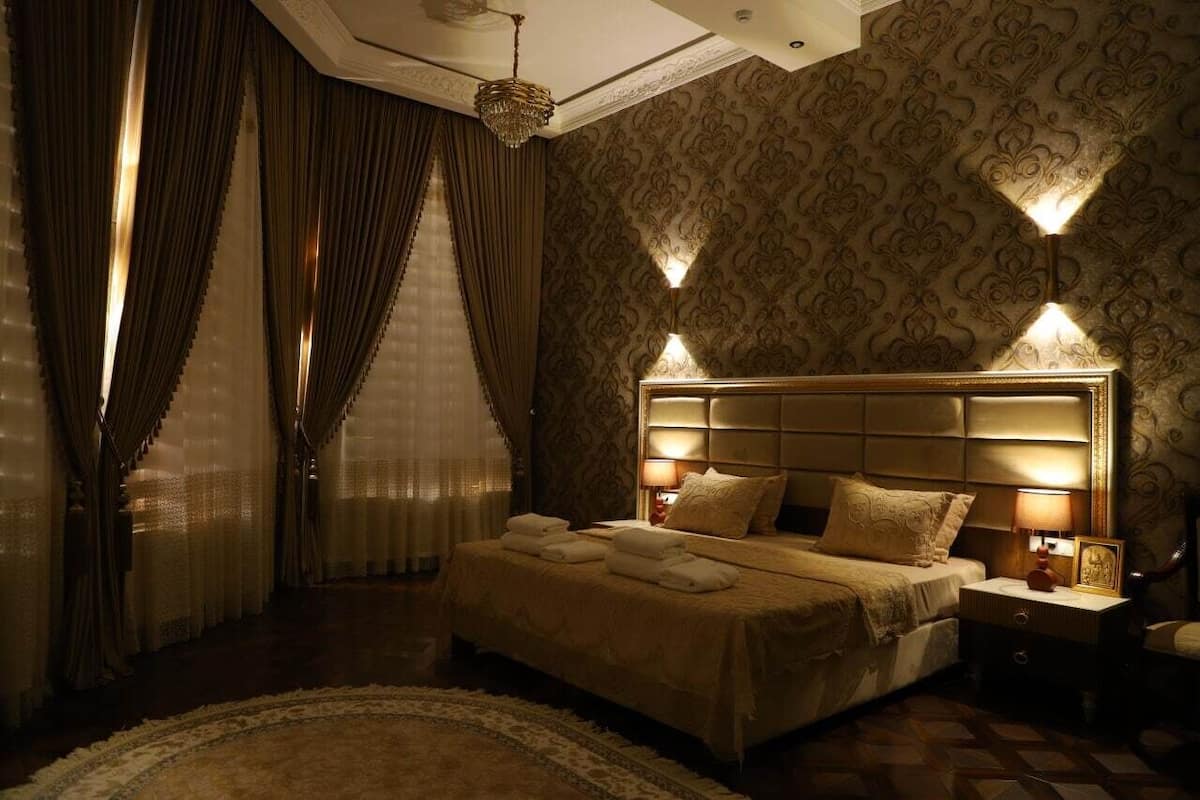 A hotel room with a large bed, a nightstand with a lamp, and a chandelier.