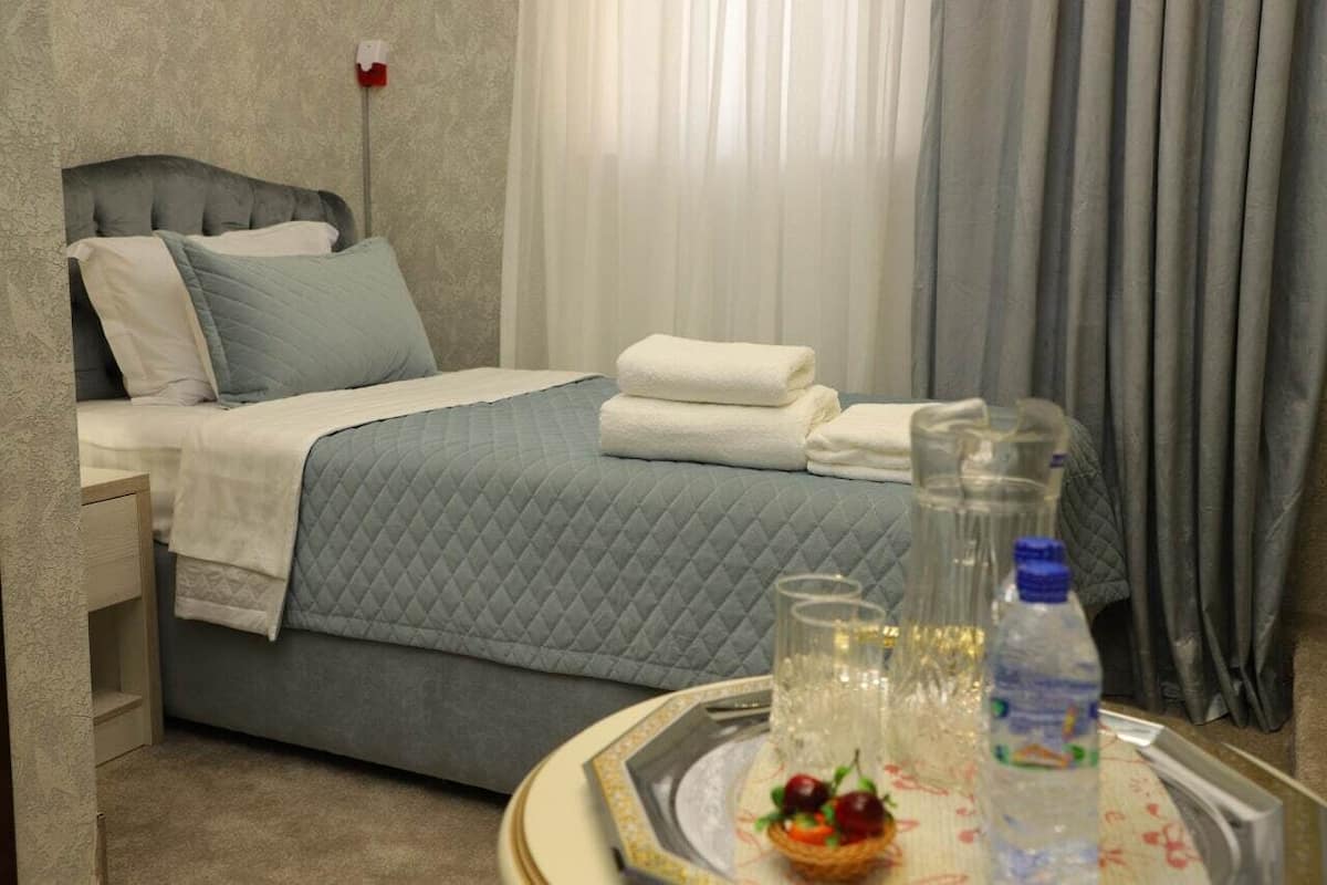 A neatly made bed with a grey quilt, white pillows, and folded towels. There is a tray with a glass of water, a bottle of water, and a small fruit arrangement.
