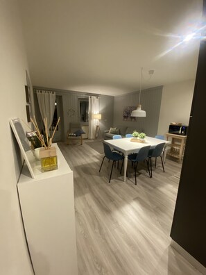 Moderni ruokailutila, jossa on valkoinen pöytä ja siniset tuolit. Pöydällä on pieni kasvi ja seinällä valkoinen kaappi, jossa on valokuvakehys ja pullo hajuvettä.
