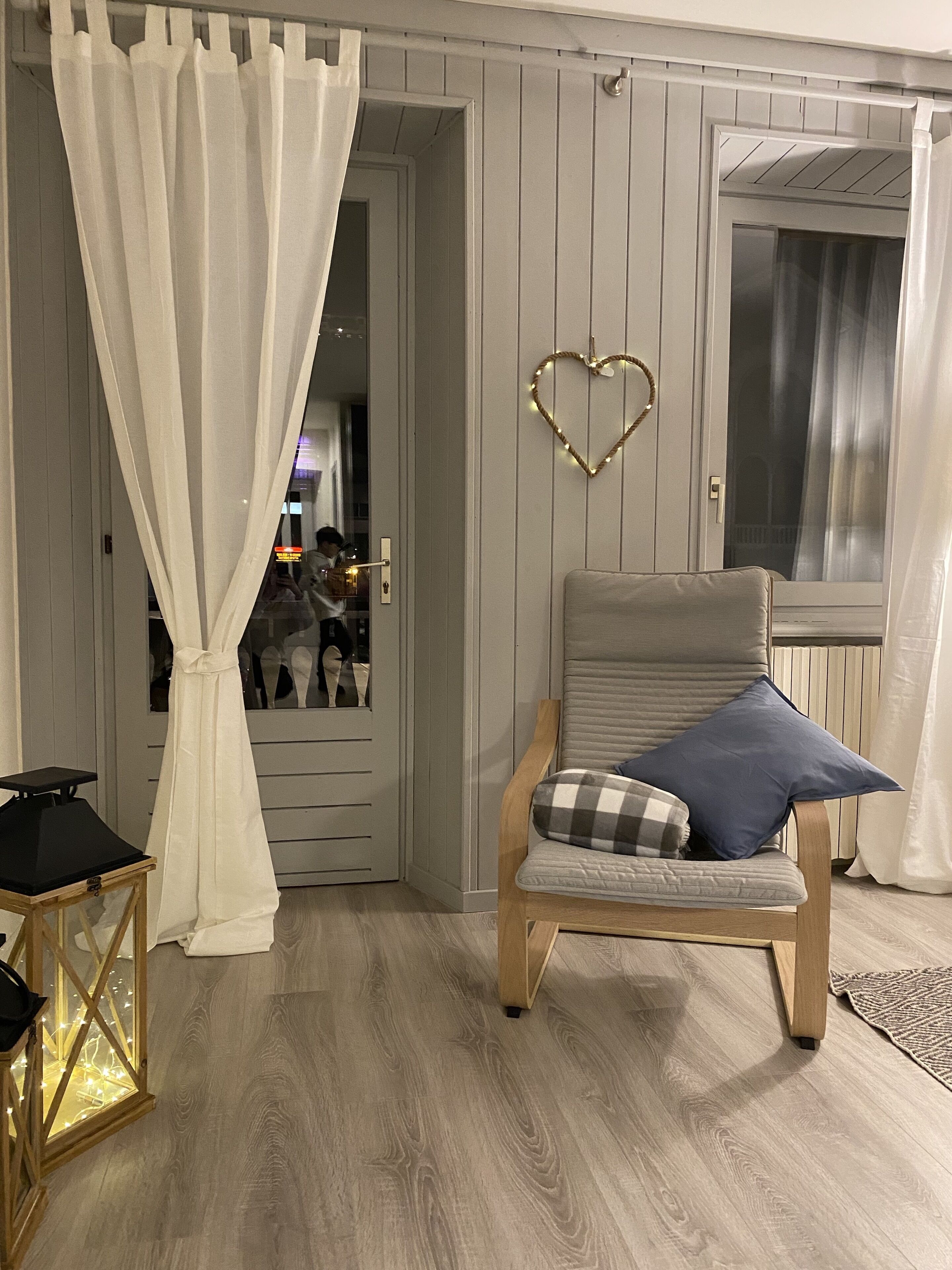Une pièce avec un plancher en bois, un fauteuil à revêtement gris agrémenté d’un coussin bleu, un rideau blanc et un luminaire en forme de cœur fixé au mur.