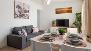 Una sala de estar moderna con un sofá gris, una mesa de comedor preparada para cuatro personas y un televisor de pantalla plana sobre un mueble.