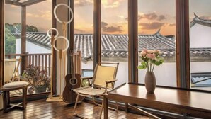 Una habitación con ventanas amplias, piso de madera, una guitarra, una silla, un jarrón con flores y una planta.