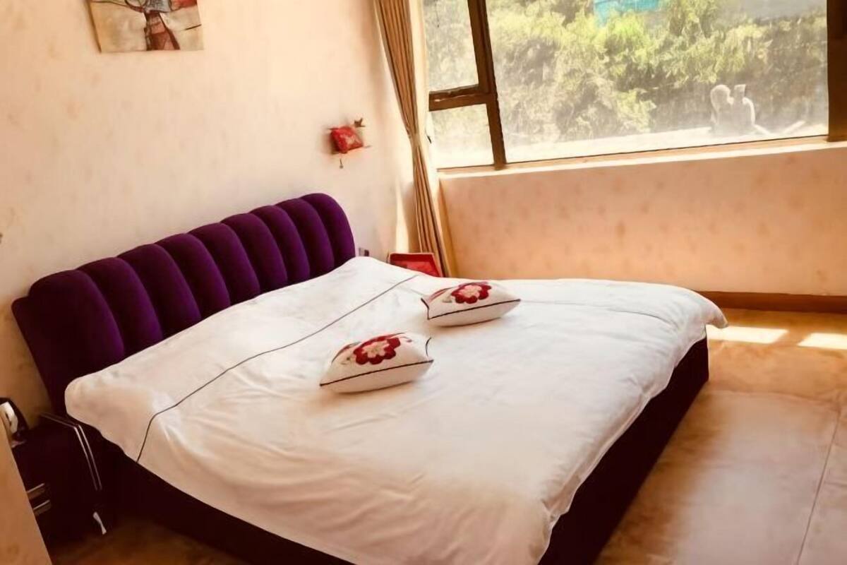 A neatly made bed with a purple headboard and white bedding, a window with a view of greenery, and a small red decoration on the wall.