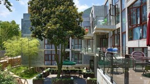 Pátio de hotel com área de estar externa, uma árvore e guarda-corpo de vidro com vista para o local.