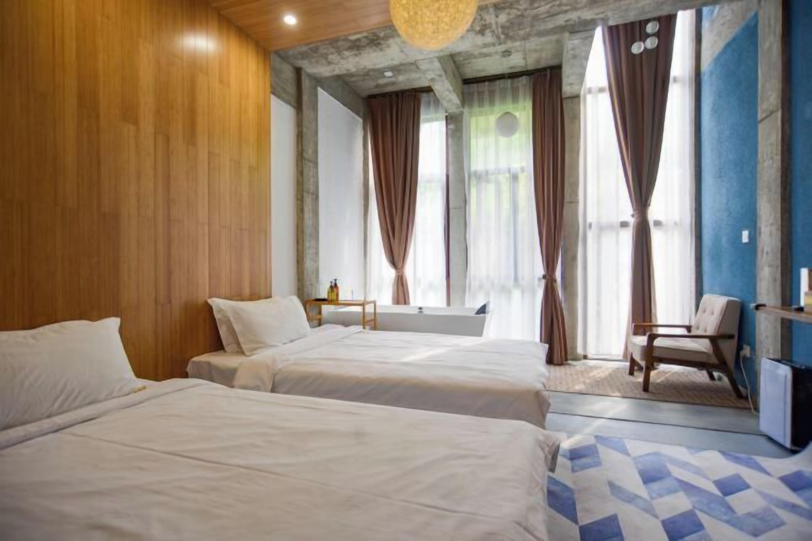 A hotel room with two beds, a wooden wall, a blue and white patterned rug, a chair, and a small table with a bottle.