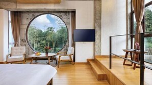 Quarto de hotel moderno com uma grande janela redonda, uma cama, uma televisão e um espaço de estar com uma mesa e cadeiras.