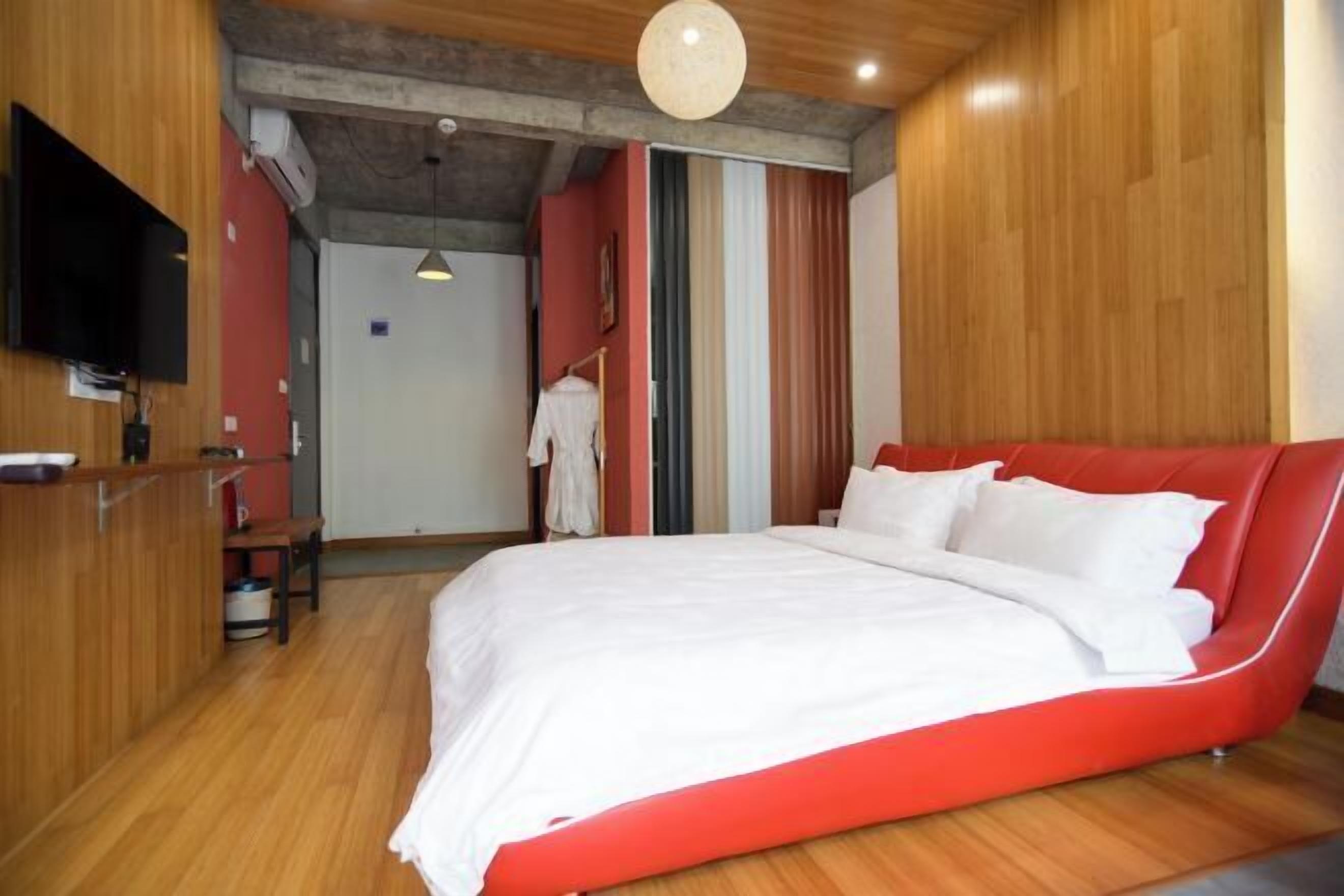 A modern bedroom with a red upholstered bed, wooden flooring, a flat-screen TV mounted on the wall, and a small desk area.