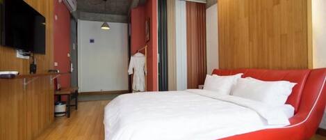 A modern bedroom with a red upholstered bed, wooden flooring, a flat-screen TV mounted on the wall, and a small desk area.