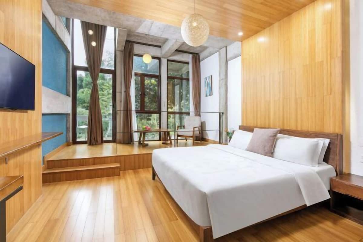 Habitación de hotel moderna con una cama grande, piso de madera, televisión y balcón con vistas a zonas verdes.