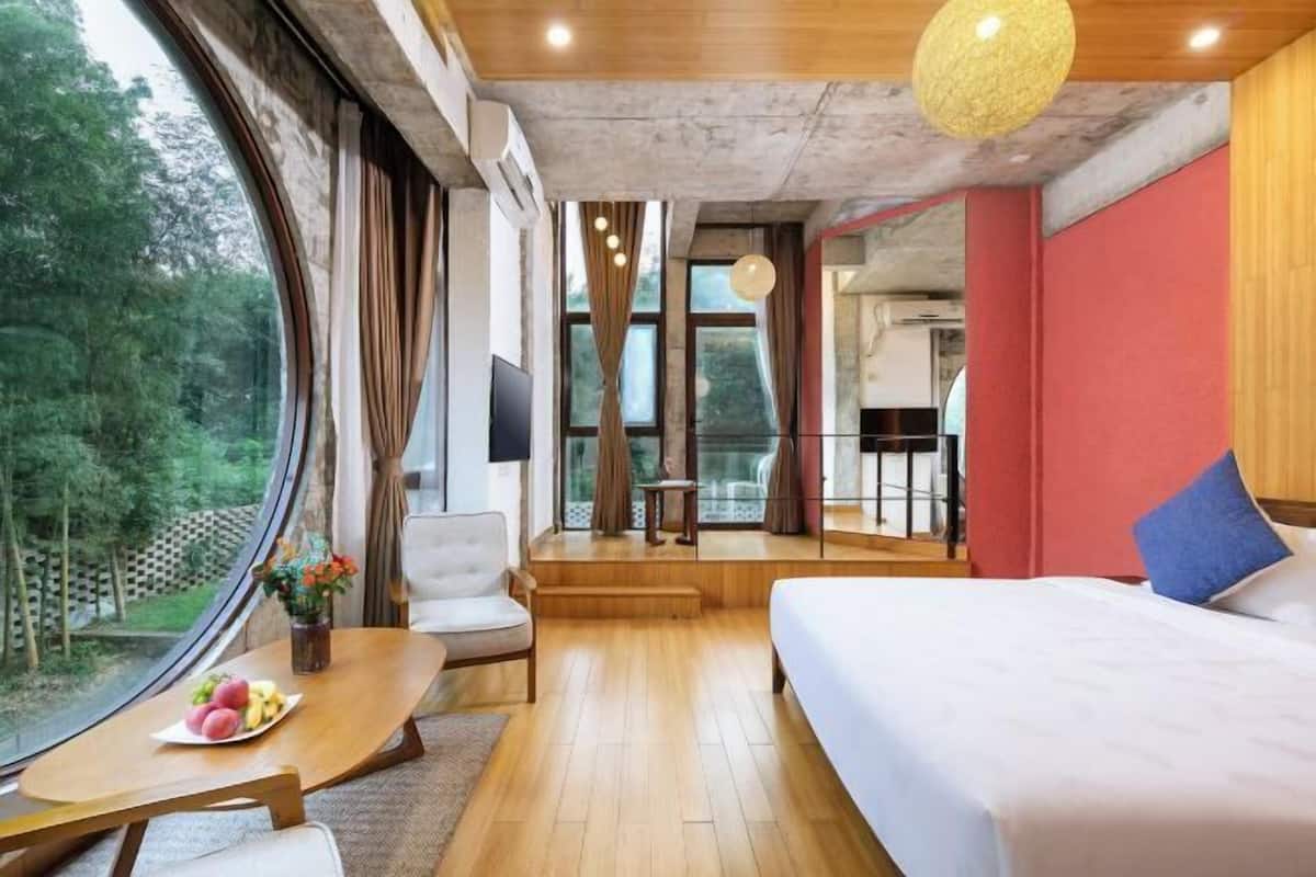 Habitación de hotel con cama, una mesa de madera con fruta, una silla y un ventanal con vistas a zonas verdes.