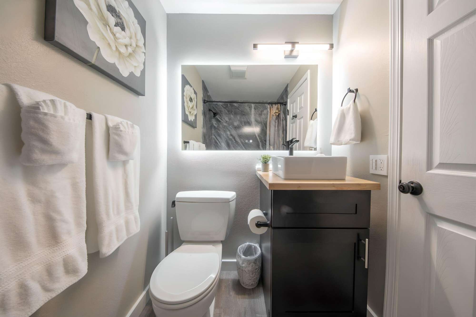 A modern bathroom with a toilet, sink, and mirror. There are towels hanging on the wall and a trash can.