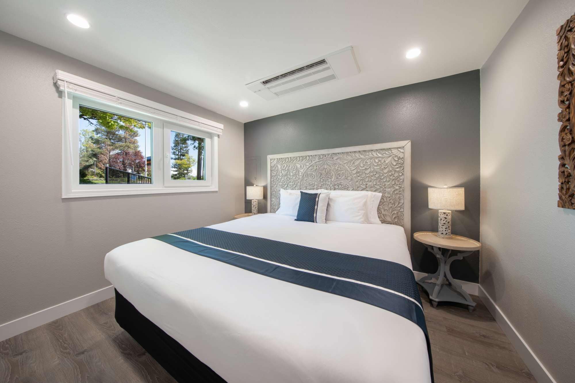 A hotel room with a large bed, a window with a view of trees, a decorative headboard, and a bedside table with a lamp.