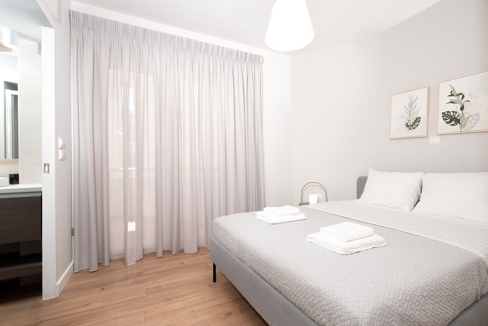 A modern hotel room with a large bed, a bedside table, a mirror, and a window with sheer curtains.