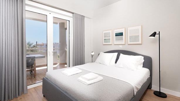 A modern bedroom with a large bed, a balcony with curtains, and a city view. - Blue Avra by A&D Properties (Markopoulo Mesogaias)