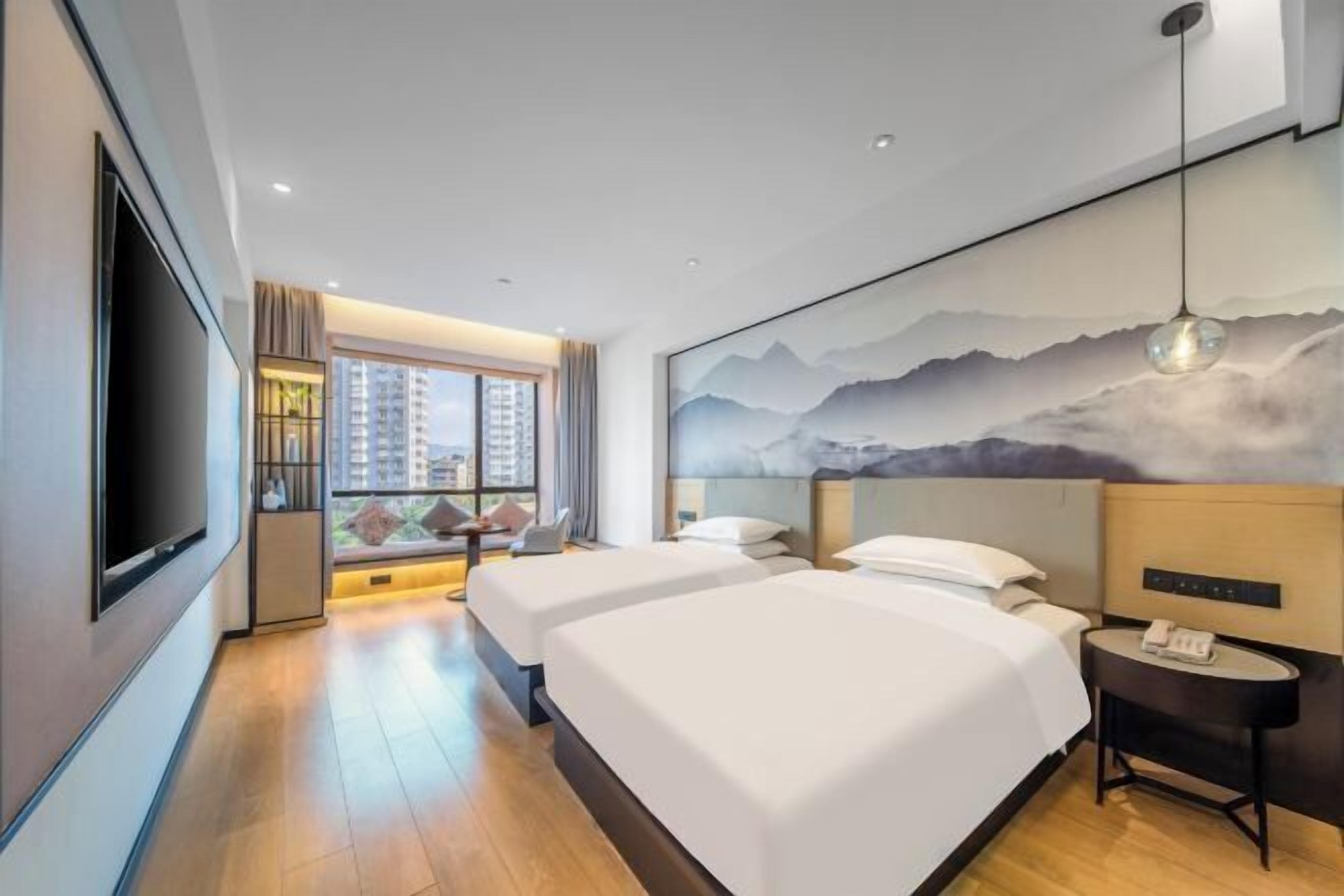 A hotel room with two beds, a TV, a desk with a phone, and a large window with a view of buildings.
