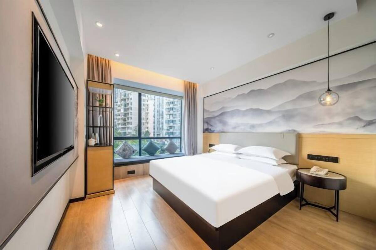 A modern hotel room with a large bed, a TV mounted on the wall, a window with a view of high-rise buildings, and a decorative wall mural.