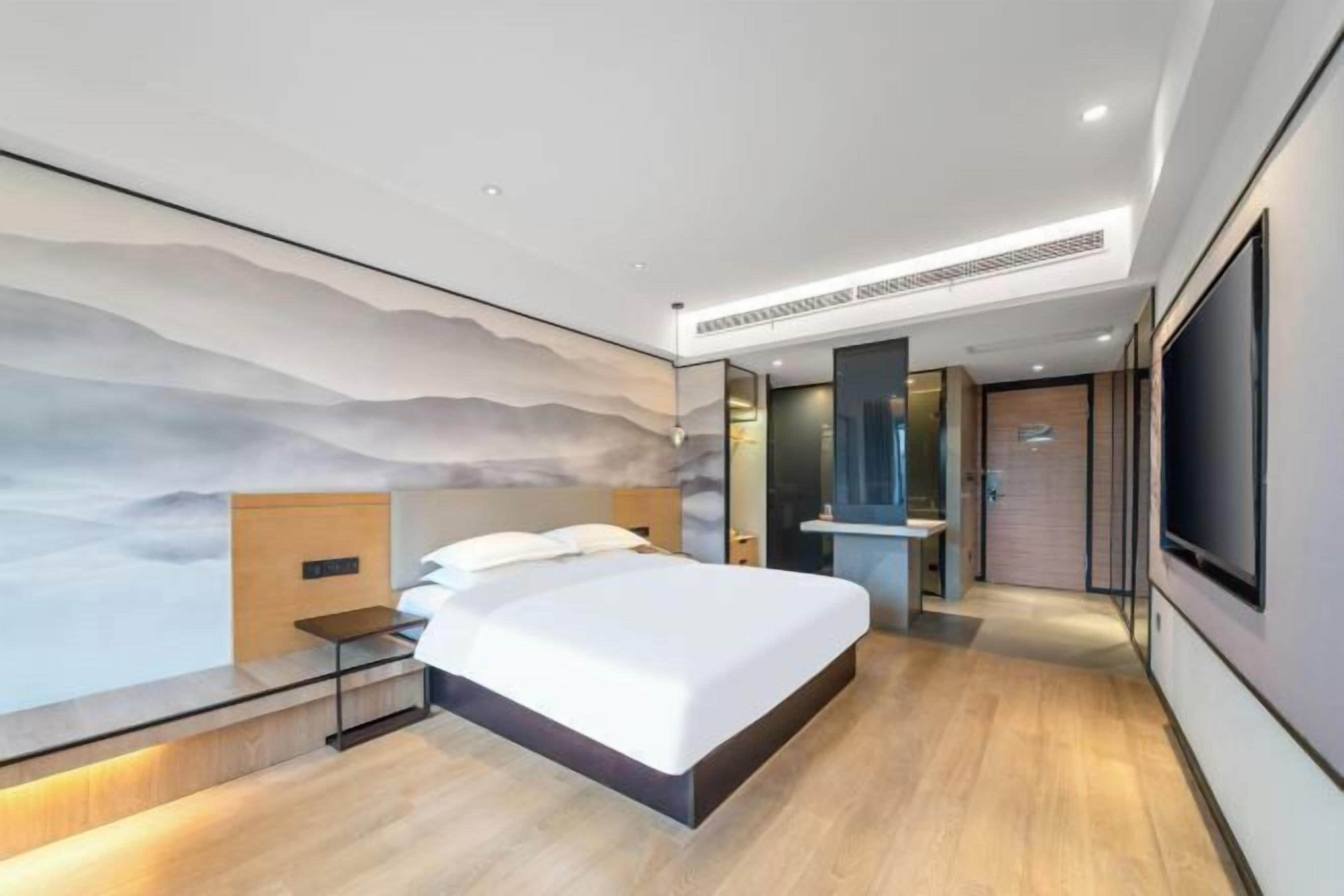 A modern hotel room with a large bed, a nightstand, a wall-mounted TV, and a bathroom visible through an open door.