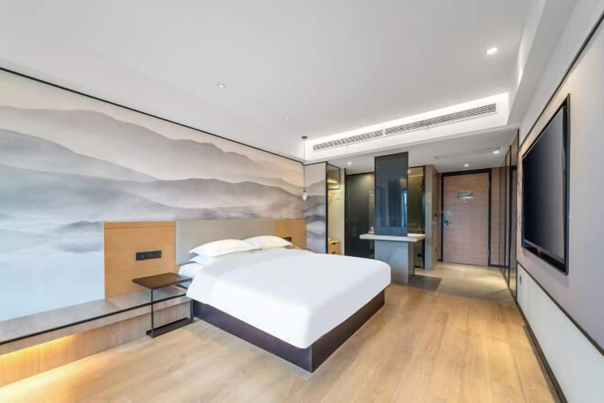 A modern hotel room with a large bed, a nightstand, a wall-mounted TV, and a bathroom visible through an open door.