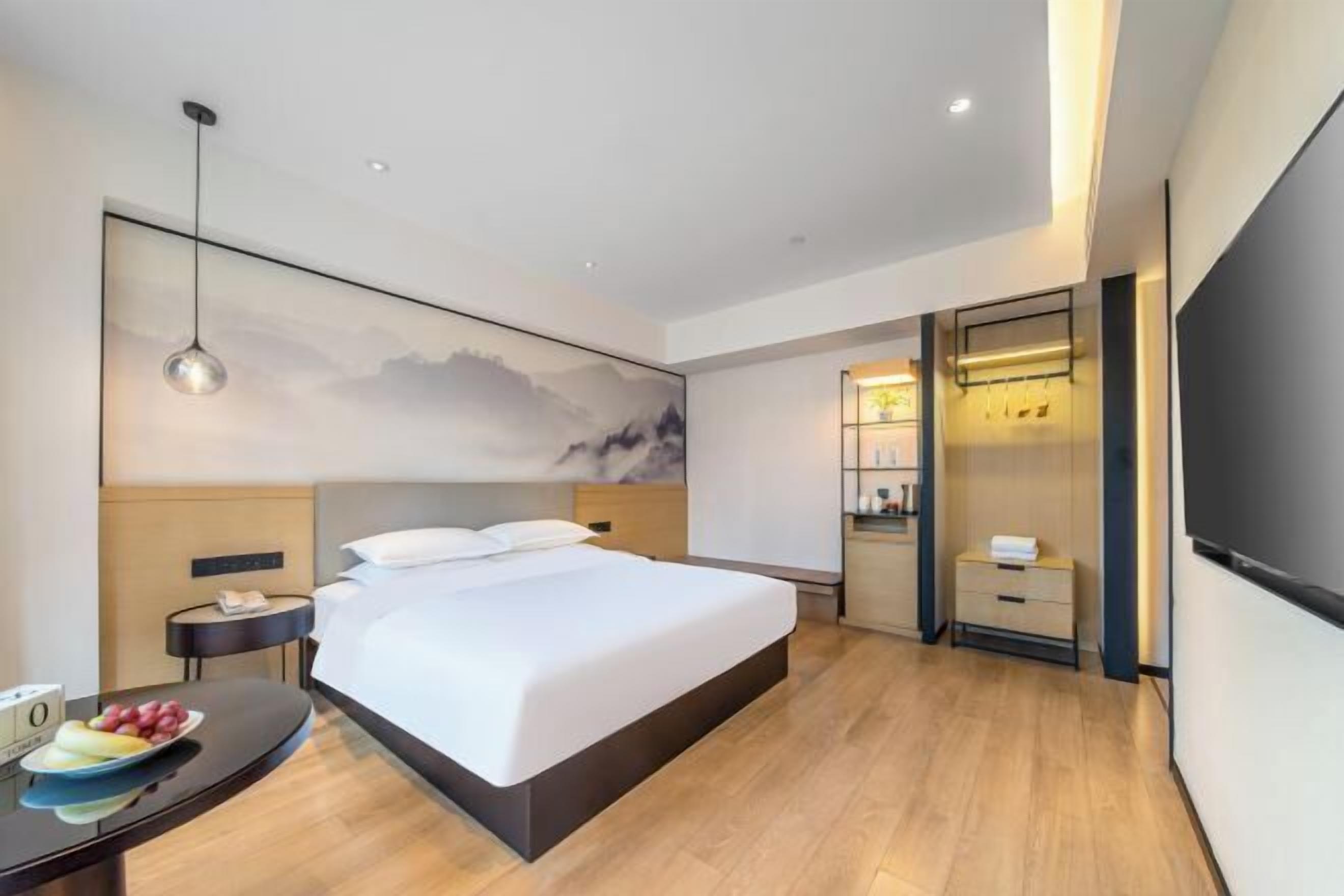 A modern hotel room with a large bed, bedside tables, a desk with fruit, a TV, and a wall-mounted shelf.