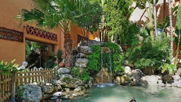 Um pátio com um pequeno lago, cerca de madeira e um restaurante chamado 'Restaurante La Lucianda'.