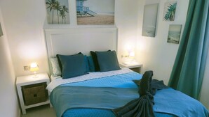 Uma cama bem arrumada com um cobre-leito estampado em azul e branco, duas luminárias de mesa e uma parede com quadros de palmeiras e praia.