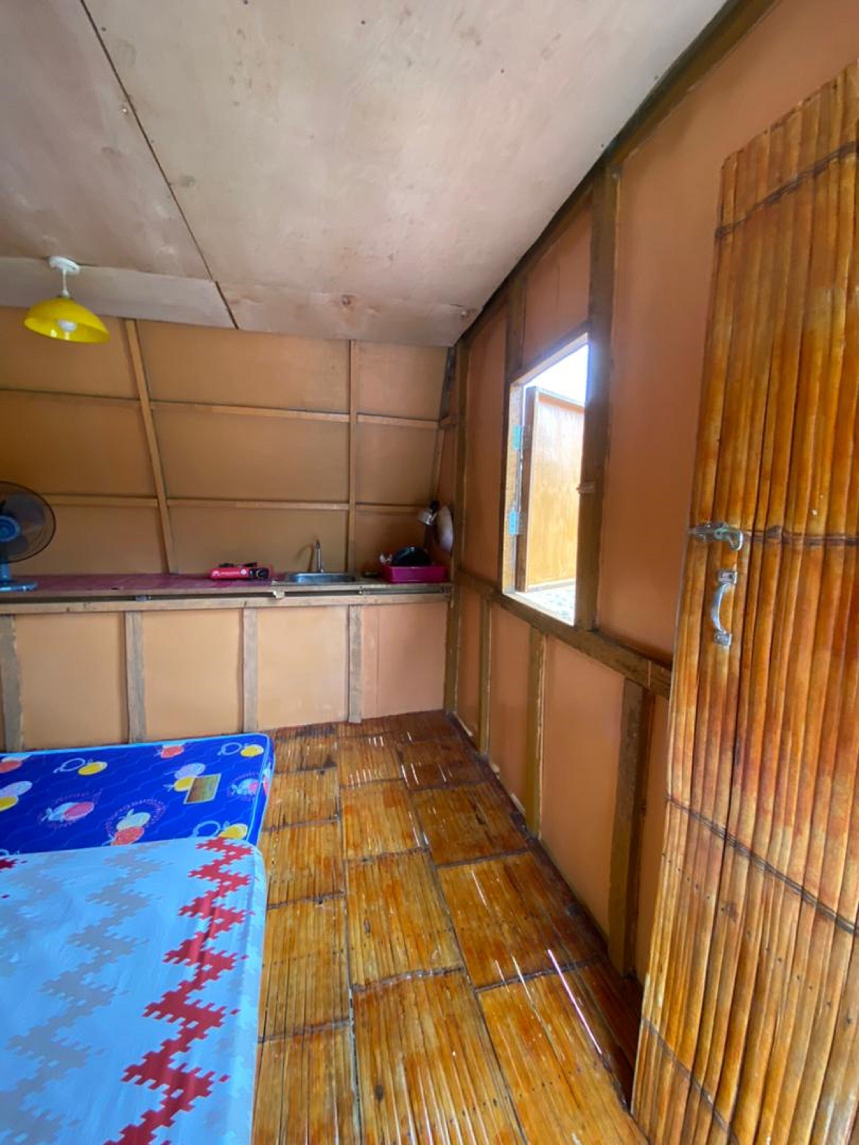 A room with a bamboo floor, a bed with a blue and red patterned bedspread, a window with a view, and a fan on the ceiling.