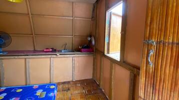 Una habitación con piso de bambú, una cama con colcha de rayas azules y rojas, una ventana con vista y un ventilador en el techo.