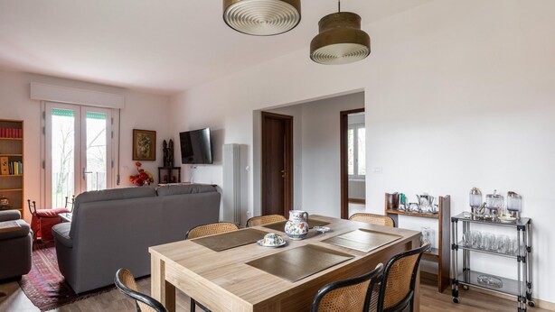 A modern living room with a sofa, dining table, and chairs. There is a TV mounted on the wall, a bookshelf, and a window with curtains. - Casa con Giardino a Borgo Panigale (Bologna)