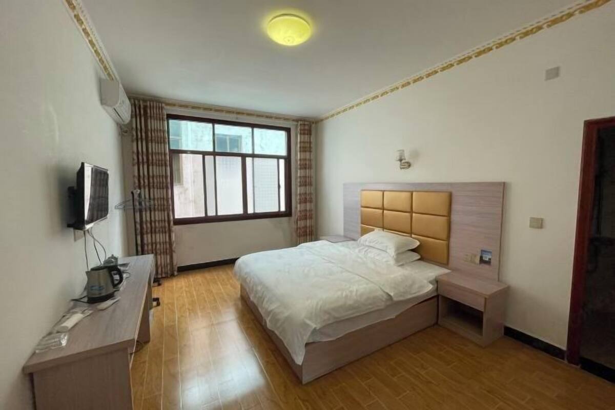 A hotel room with a bed, a desk with a kettle, a TV, and a window with curtains.