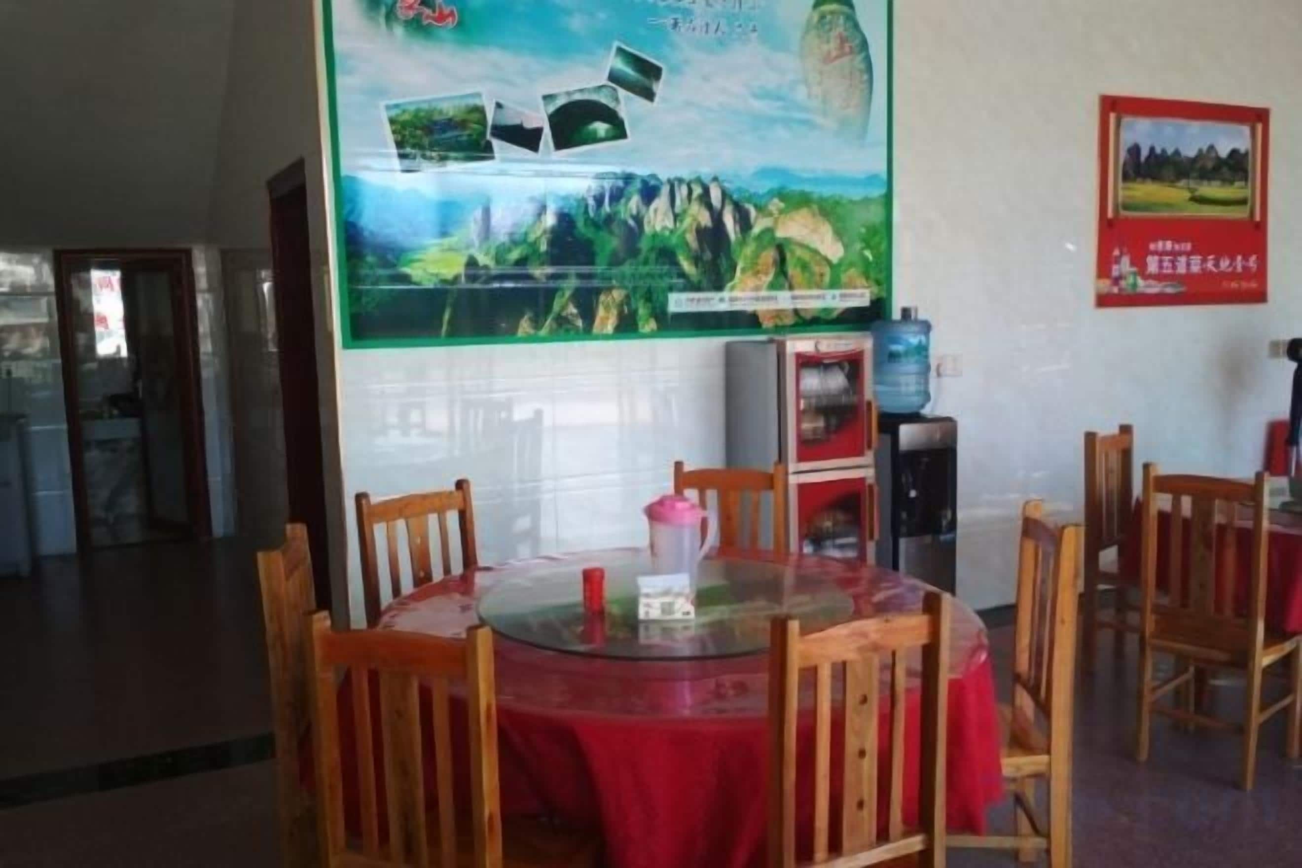 A dining area with a round table covered in a red tablecloth, surrounded by wooden chairs. There is a poster on the wall behind the table, and a water dispenser is visible to the right.