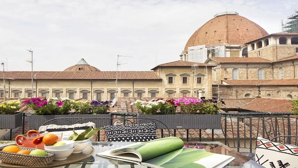 Una terraza con una mesa dispuesta para una comida, un libro y un cojín decorativo. Al fondo, un edificio con una cúpula y un campanario.