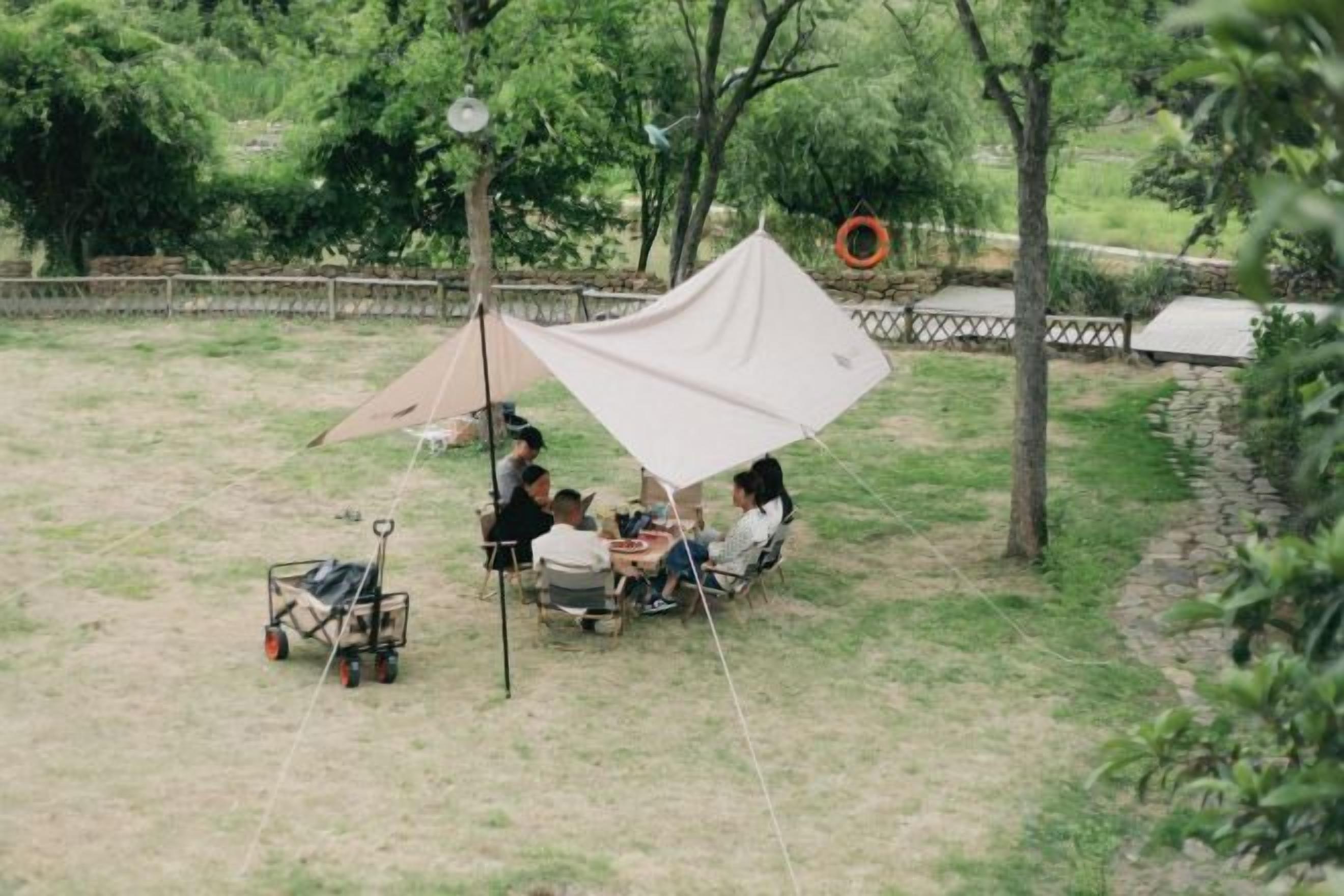 A group of people sitting at a table under a large tent in an outdoor setting with trees and a stone wall in the background.
