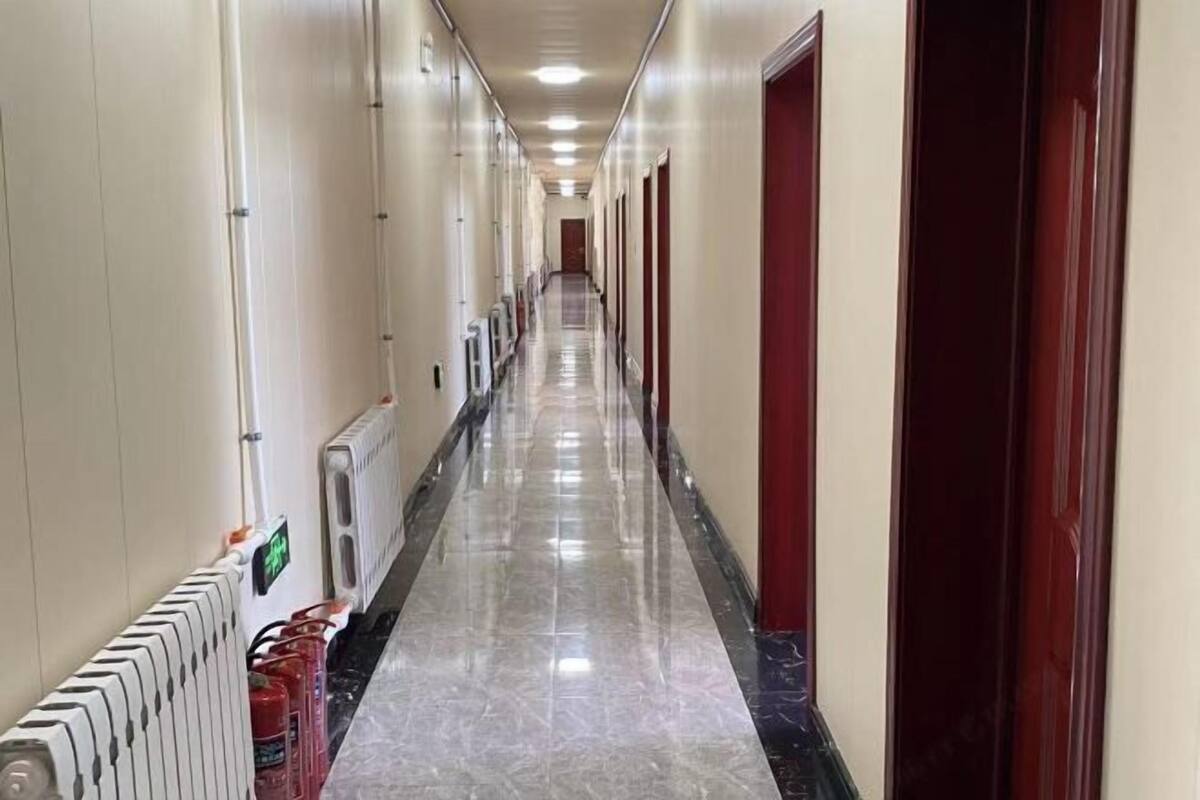 A long, narrow hotel corridor with doors on both sides, a radiator on the left, and a fire extinguisher on the right.