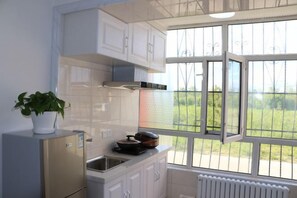 A kitchen with white cabinets, a stainless steel refrigerator, a window with a view of greenery, and a potted plant on the counter. - Tahe Eighteen Post Residence (Tahe)