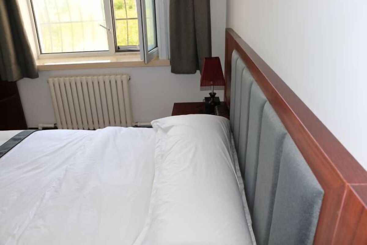 Una habitación de hotel con una cama, una ventana con cortinas, una mesita de noche con lámpara y un radiador.