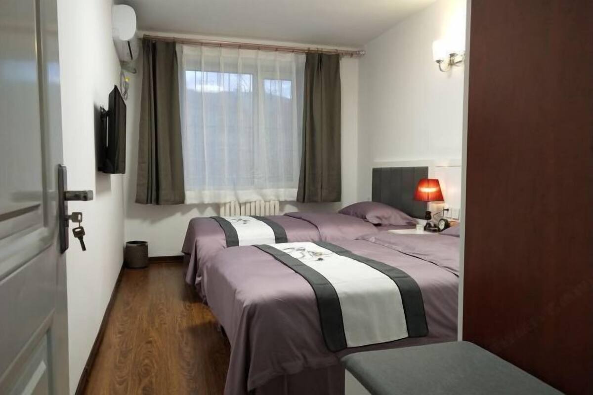 A bedroom with two beds, a window with curtains, a bedside lamp, and a radiator.