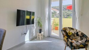 A room with a flat-screen TV mounted on the wall, a potted plant, a floral-patterned chair, and a sliding glass door leading to an outdoor area with a red umbrella.