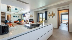 A modern kitchen with a central island, a dining area with a table and chairs, and a view of the sea through large windows. - Lusol Seafront Villas (Chania)