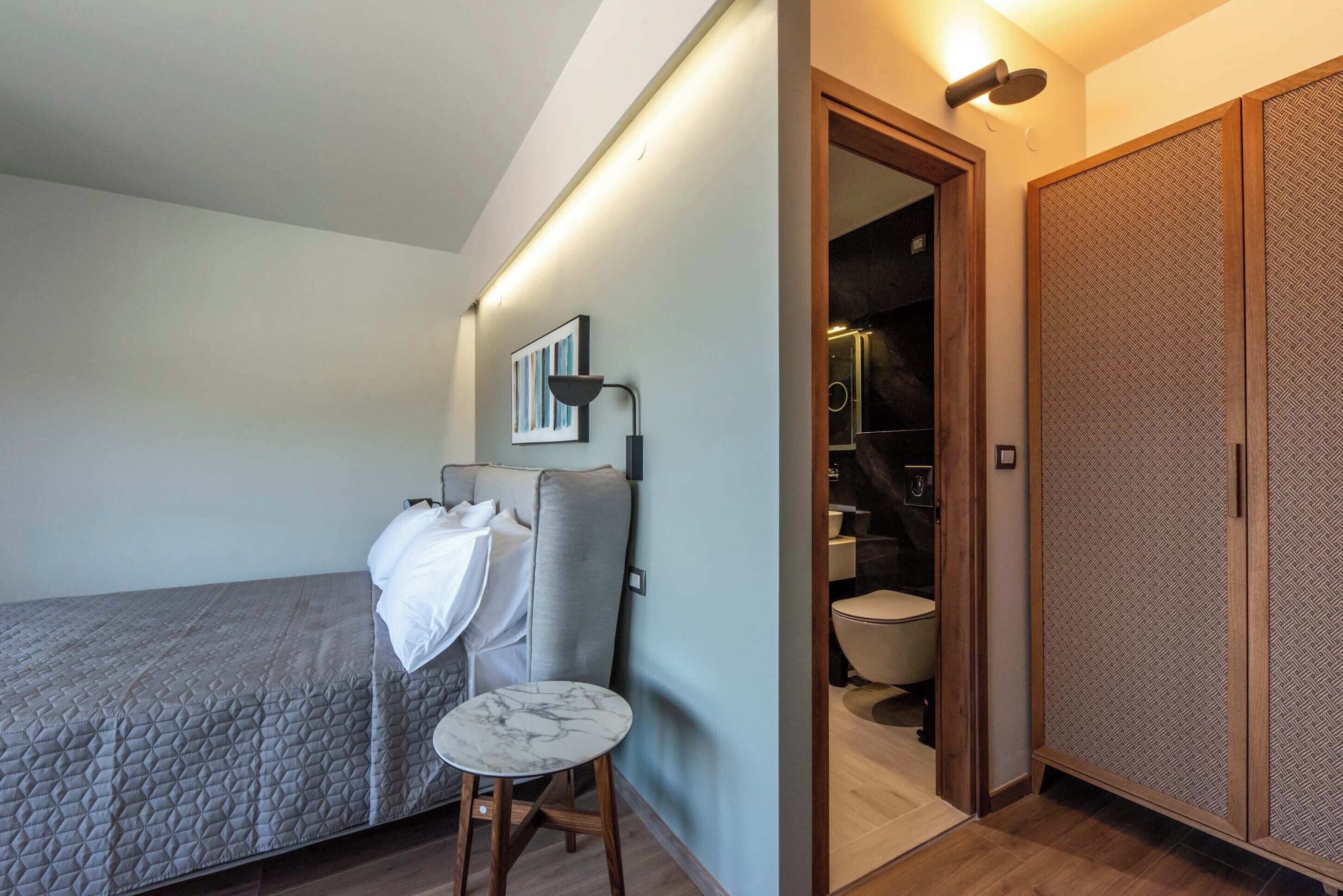A bedroom with a bed, a small table, a chair, a bedside lamp, and a wardrobe.