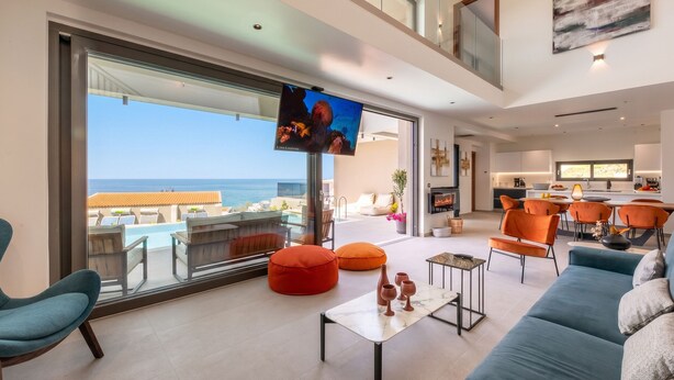 A modern living room with a large TV, comfortable seating, a kitchen area, and a view of the ocean. - Lusol Seafront Villas (Chania)