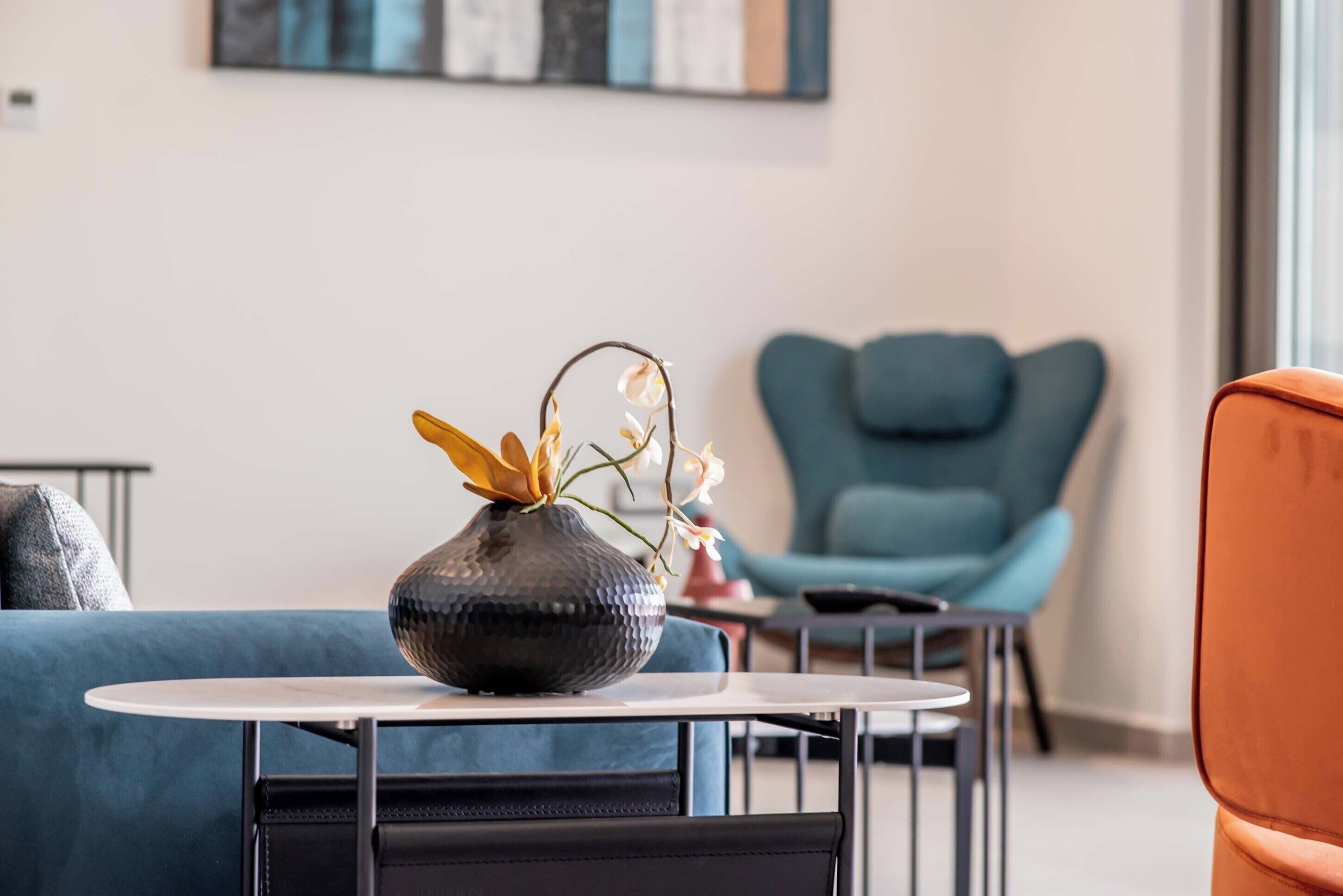 A modern living room with a round table, a dark vase with flowers, a blue sofa, and a teal armchair.