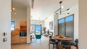 A modern kitchen with wooden cabinets, a dining area with a round table and chairs, and a view through a large window. - Lusol Seafront Villas (Chania)