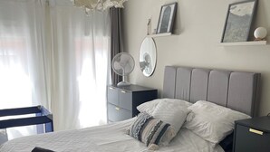 A neatly made bed with a white quilt, pillows, and a rolled towel. There is a bedside table with a fan, a wall-mounted shelf with pictures, and a large, ruffled light fixture above the bed.