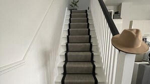 A staircase with carpeted steps and a black handrail, leading to a landing with a framed picture and a plant.